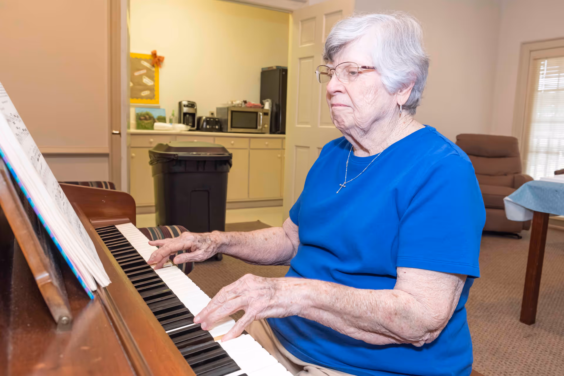 An elderly woman with short gray hair and glasses is playing a piano in a room. She is wearing a blue shirt and a cross necklace. In the background, there is a kitchenette area with a microwave, coffee maker, and a black trash bin. The room also contains a brown recliner chair and a table with a blue tablecloth near a window with blinds.
