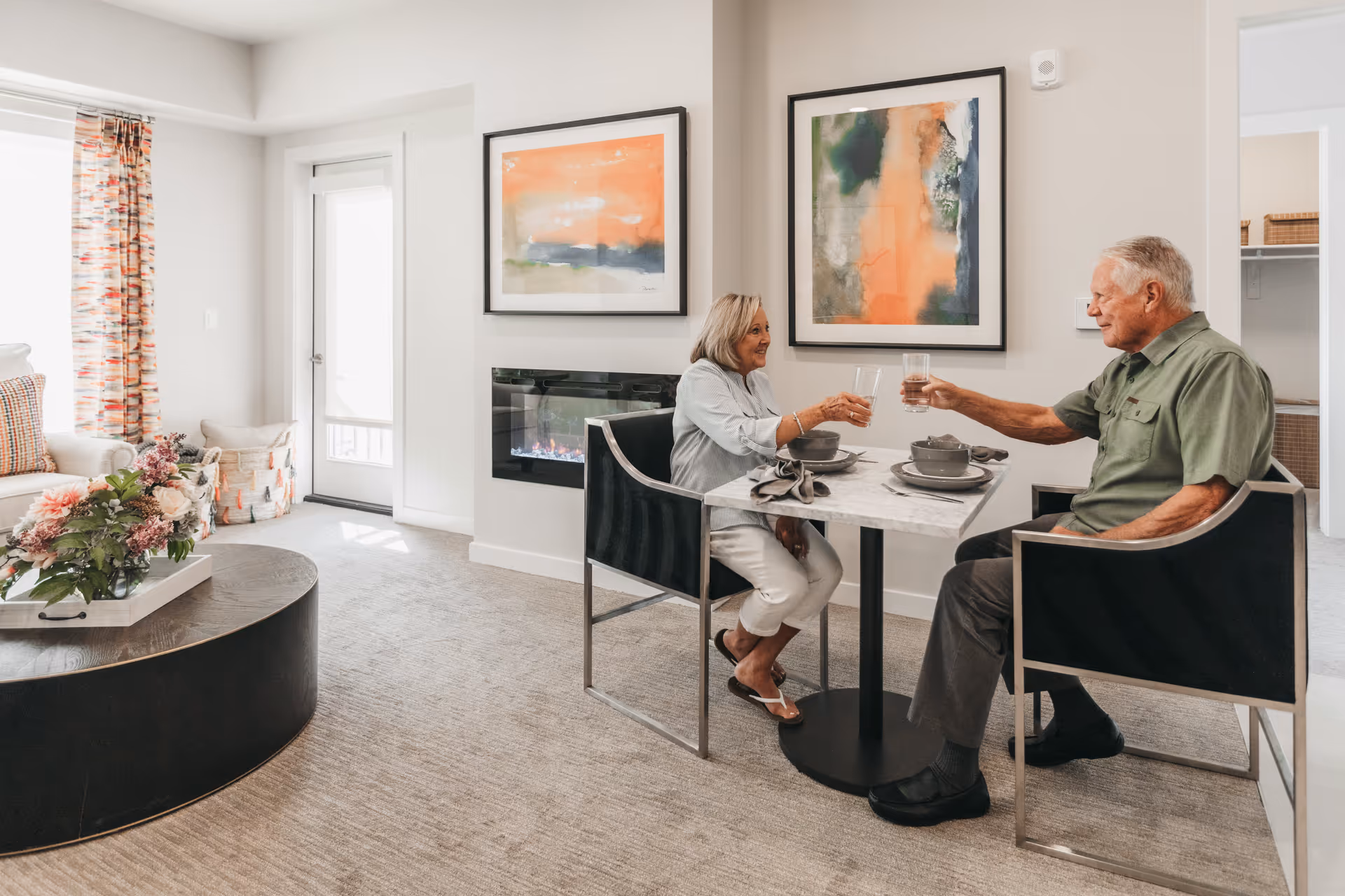 An elderly couple sitting at a small dining table in a cozy living room area, clinking glasses of water. The room features a modern electric fireplace, abstract artwork on the walls, a round coffee table with a floral arrangement, and a door with patterned curtains letting in natural light.