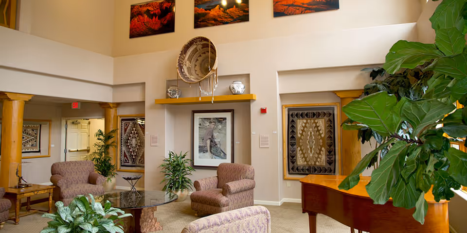 A spacious and well-lit living room area in a senior living community featuring comfortable upholstered armchairs arranged around a glass coffee table, large green plants, a wooden piano, and decorative Native American artwork and pottery displayed on the walls and shelves.