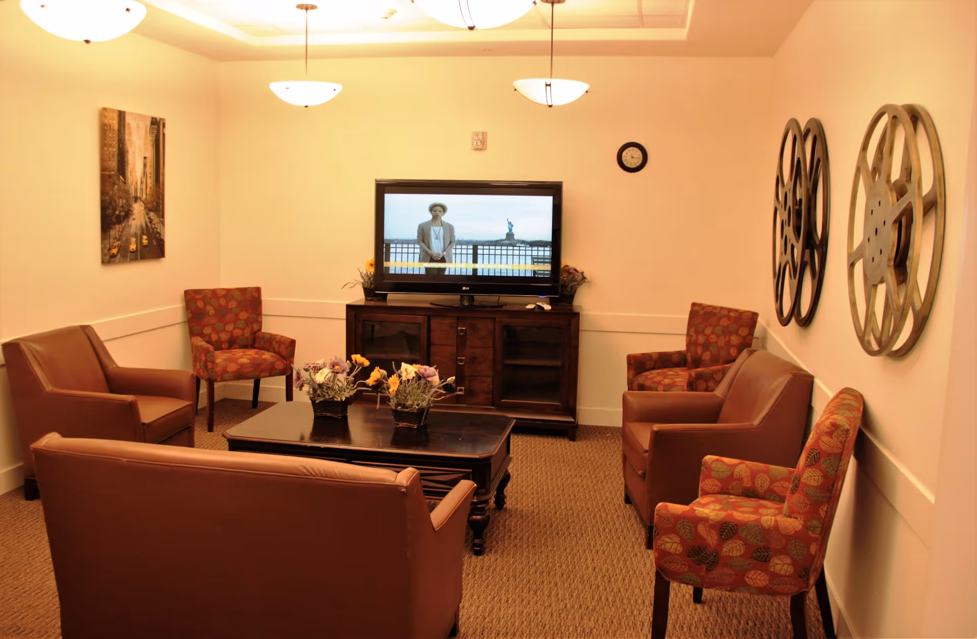 A cozy living room with a brown leather loveseat and two matching armchairs, along with three patterned fabric chairs arranged around a dark wooden coffee table with flower arrangements. A flat-screen TV is mounted on a wooden cabinet against the wall, showing a person standing near the Statue of Liberty. The walls are decorated with two large film reel wall hangings and a framed picture of a city street with yellow taxis. The room is softly lit by ceiling lights.