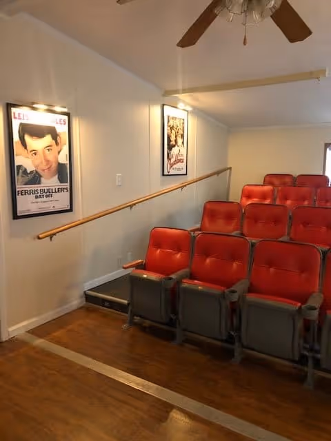 A small theater room with two rows of red cushioned seats facing a blank wall. The walls are light-colored with two movie posters framed and lit on the left wall. There is a wooden handrail along the wall and a ceiling fan with wooden blades above.
