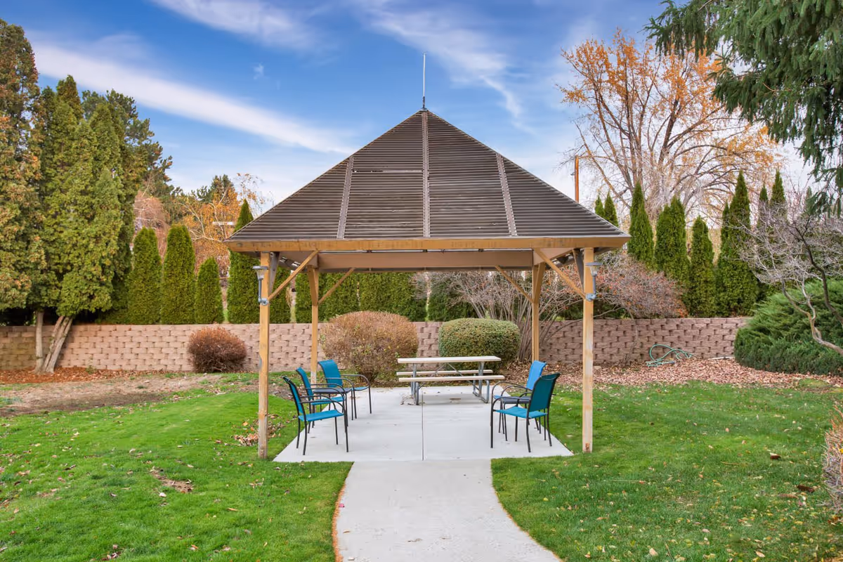 Outdoor gazebo structure with a wooden roof and six blue chairs arranged around a picnic table on a concrete slab, surrounded by green grass, bushes, and tall trees under a partly cloudy sky.