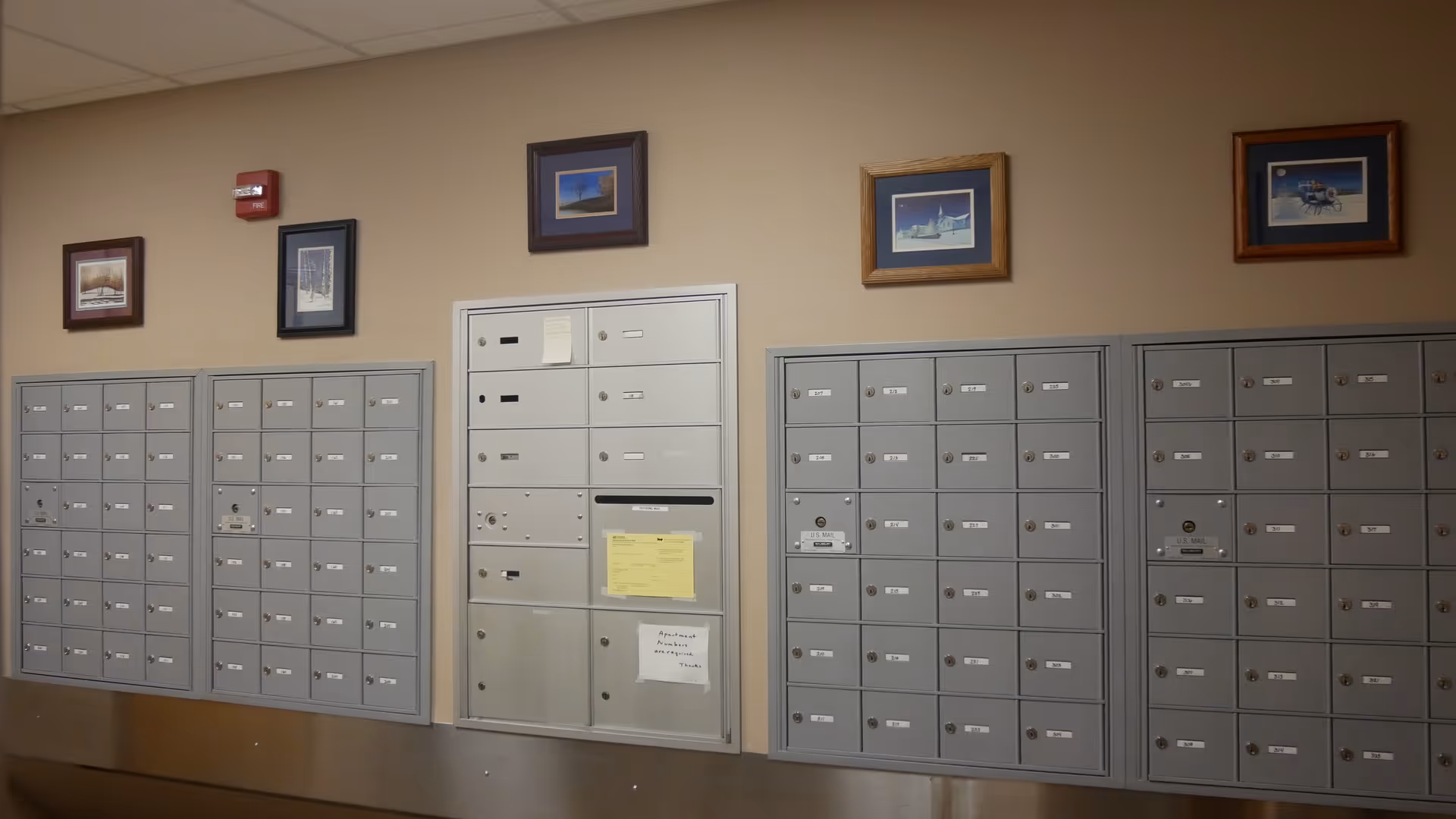 A wall with multiple gray mailboxes arranged in rows and columns, with four framed pictures hanging above them on a beige wall. There is a fire alarm on the wall to the left.