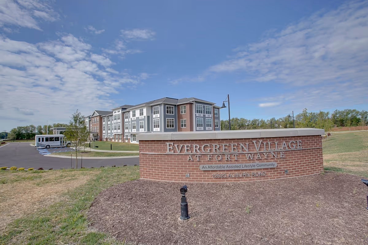 Brick entrance sign reading "Evergreen Village at Fort Wayne" in front of a multi-story senior living building and shuttle under a partly cloudy sky.