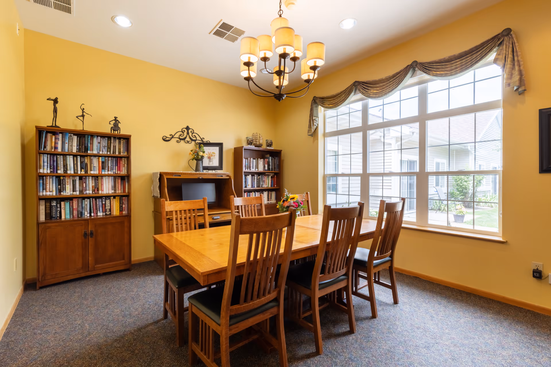 Sunlit dining room with a wooden table and chairs, bookshelves, and a large window.