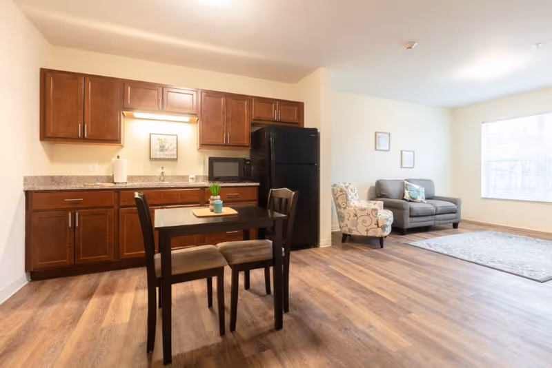 A bright and clean living space featuring a small kitchen area with dark wooden cabinets, a black refrigerator, microwave, and a countertop with a paper towel holder. In front of the kitchen is a small dining table with two chairs. To the right, there is a cozy sitting area with a patterned armchair, a gray sofa with decorative pillows, and a large window letting in natural light. The floor is covered with wood-style laminate.