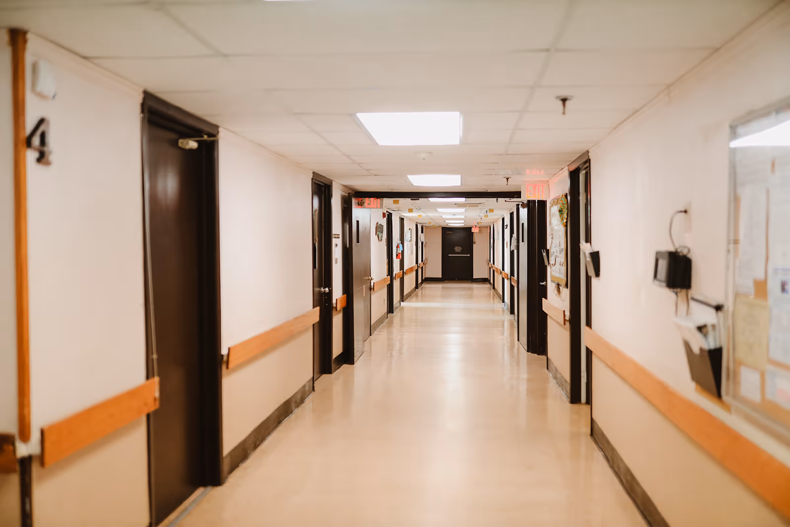 A long, clean hallway in a healthcare facility with beige walls, wooden handrails, and multiple closed dark brown doors on both sides. The ceiling has recessed lighting panels, and there are exit signs visible at the far end of the corridor.