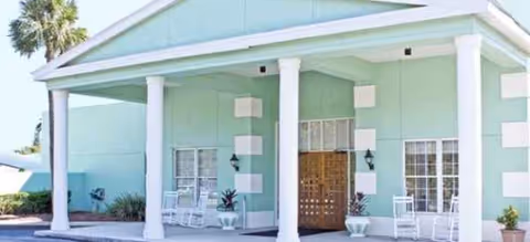 Mint-green senior living building entrance with columns, benches, and potted plants.