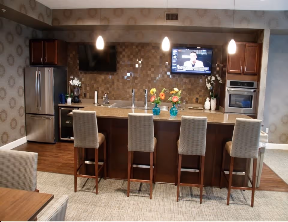 Modern kitchen area with a large island featuring four high chairs. The kitchen has stainless steel appliances including a refrigerator and built-in oven. Two flat-screen TVs are mounted on the wall above the countertop, which is decorated with two blue vases holding colorful flowers. The backsplash has a textured tile design, and the room has pendant lighting and patterned wallpaper.