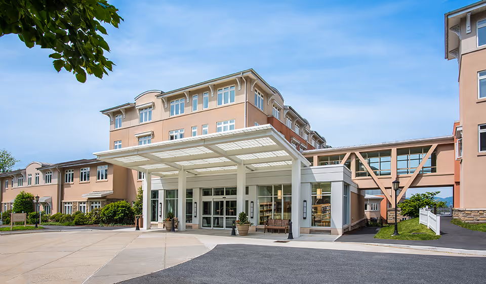 Exterior view of Kendal On Hudson senior living facility showing the main entrance with a covered drop-off area, multiple windows, and a sky bridge connecting two buildings under a clear blue sky.