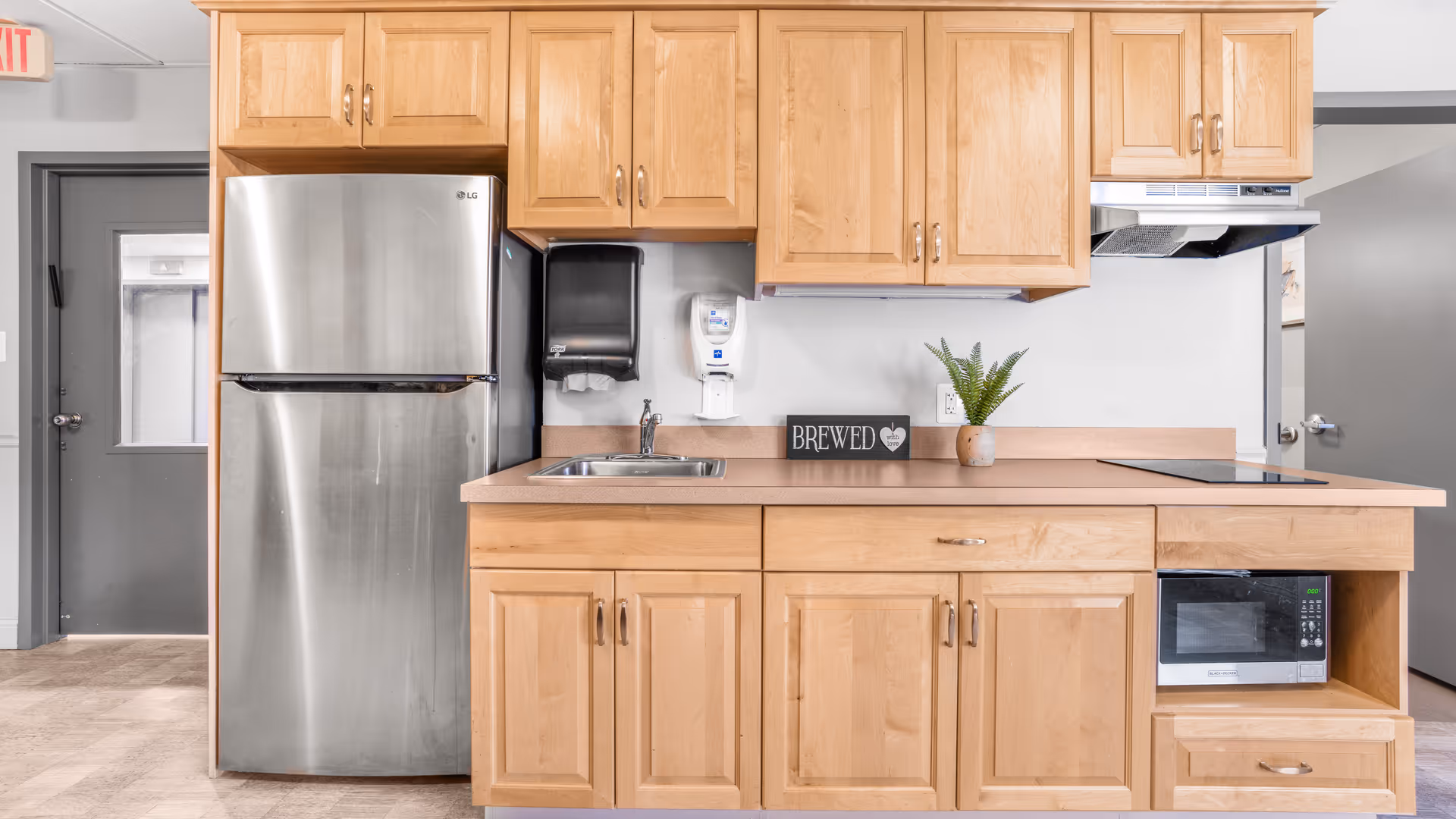 A clean kitchen area with light wood cabinets, a stainless steel refrigerator, a sink, a microwave, and an electric stovetop. There is a small potted plant and a decorative sign that says 'BREWED with love' on the countertop. The kitchen is set against a white wall with a paper towel dispenser and a soap dispenser mounted above the sink. Two gray doors are visible in the background.