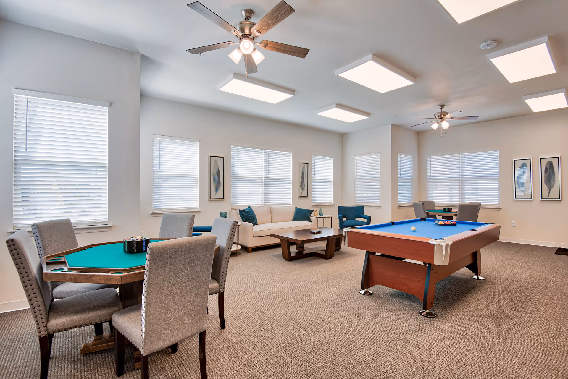 A spacious common room with a pool table and a card table with four chairs. The room has beige carpeting, multiple windows with white blinds, two ceiling fans with lights, a beige sofa with blue cushions, and framed artwork on the walls.