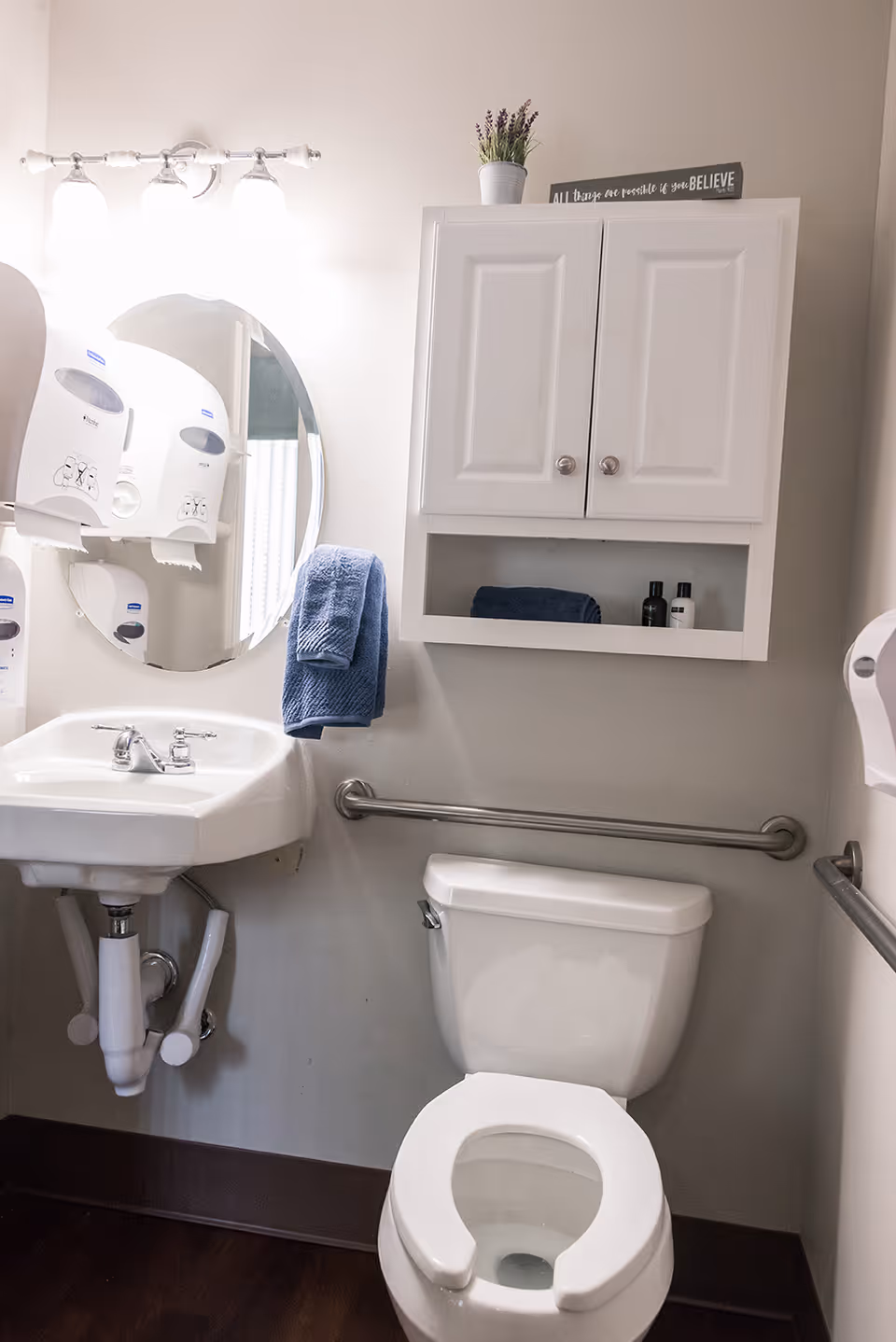 Small accessible bathroom with a toilet, wall-mounted sink and mirror, white storage cabinet, grab bars and a blue towel.
