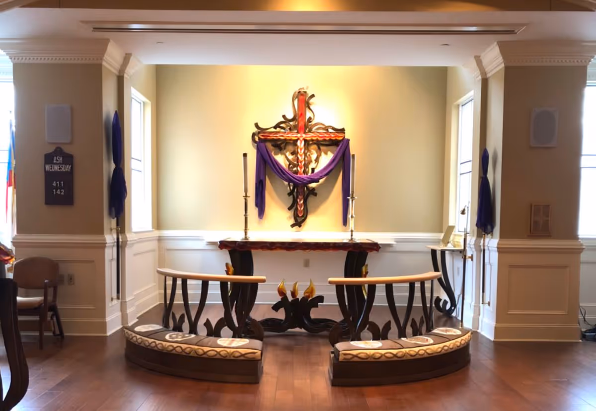 Interior view of a small chapel or prayer room with a decorative cross draped in purple cloth on the wall, two tall candlesticks on an altar table, and two curved wooden kneelers with cushioned tops in front. The room has wooden flooring, light-colored walls with white wainscoting, and windows on either side letting in natural light.