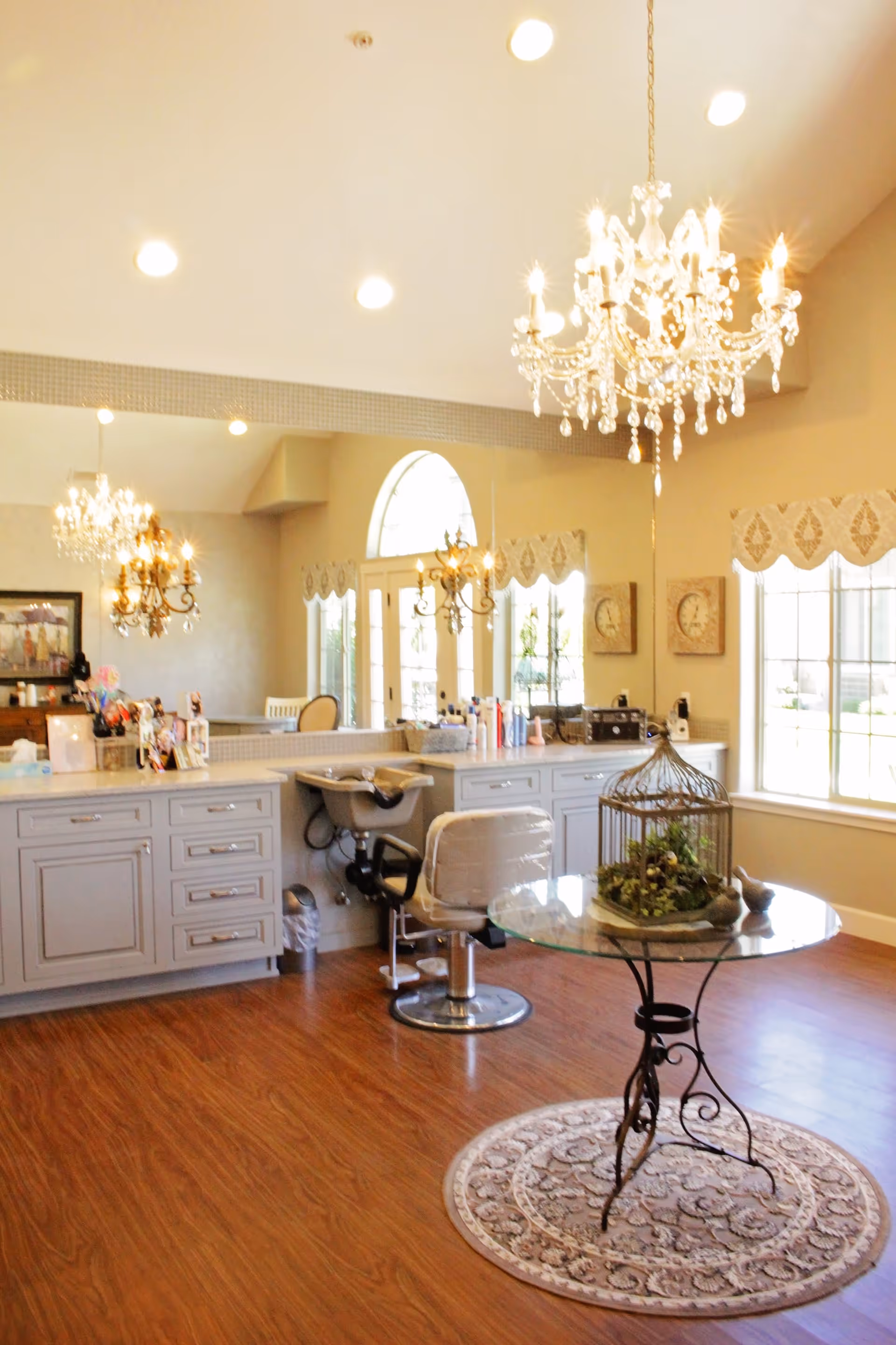 A bright and elegant salon area with wooden flooring, a round glass table with a decorative birdcage centerpiece on a patterned rug, a salon chair in front of a counter with drawers and hair care products, large windows with valances, and two ornate chandeliers hanging from the ceiling.