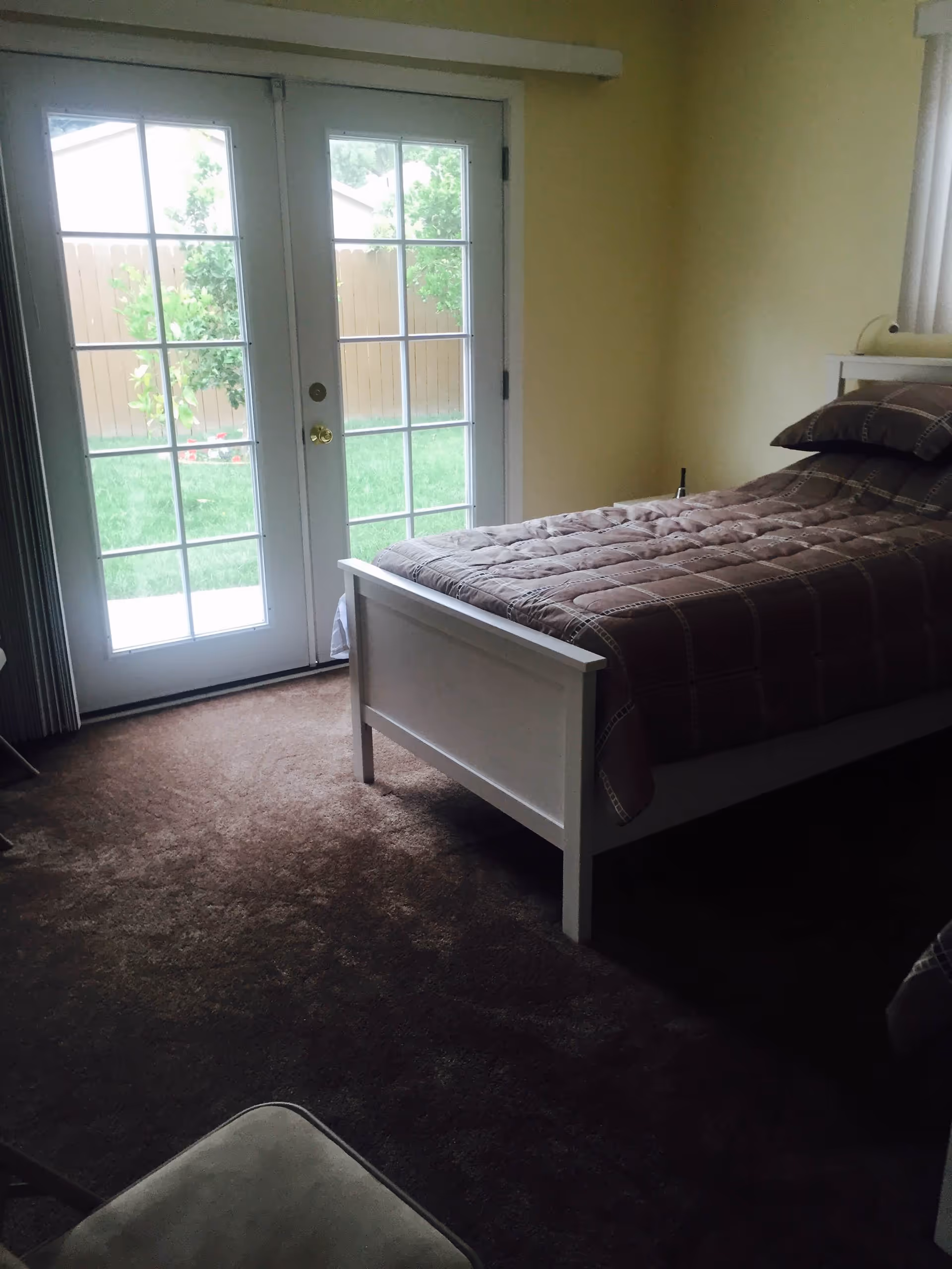 A bedroom with a single bed covered in a brown quilt and matching pillow. The room has beige walls and brown carpet. There are glass double doors with white frames leading outside to a green lawn and wooden fence.