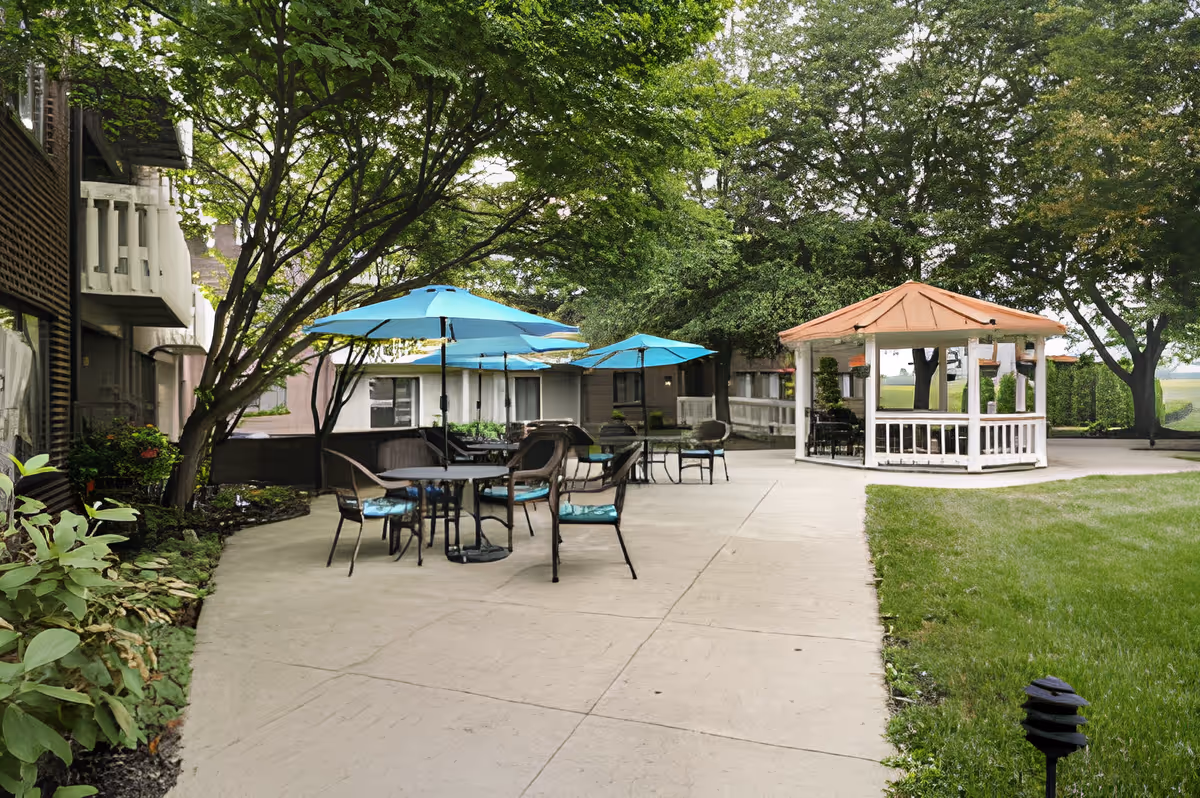 Outdoor patio area at King's Reserve College Park featuring several tables with blue umbrellas and chairs, surrounded by trees and greenery. A white gazebo with a brown roof is visible on the right side, and the area is paved with concrete walkways.