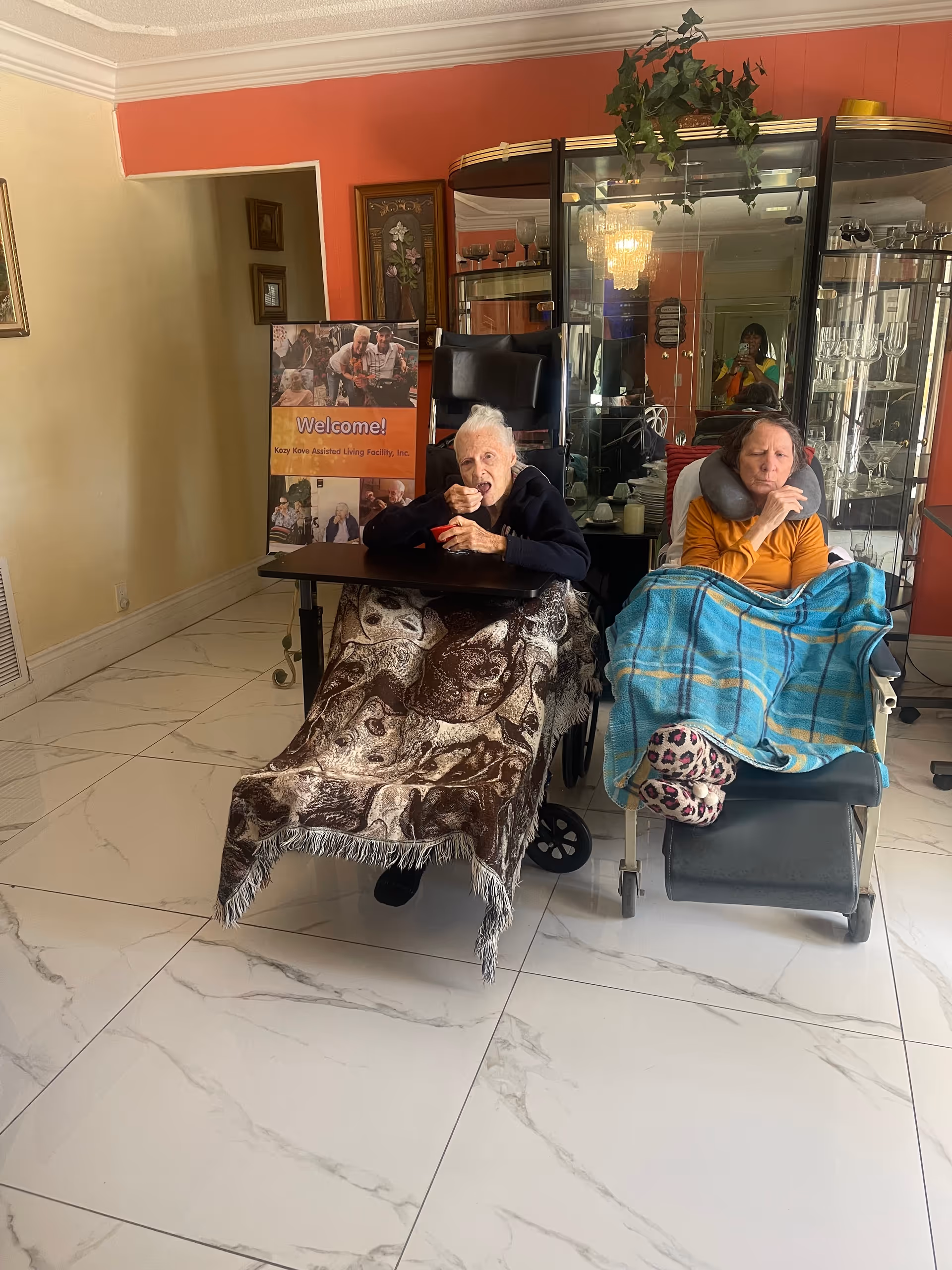 Two elderly women sitting in wheelchairs inside a room with white tiled floor and red walls. One woman is eating and covered with a brown patterned blanket, while the other is wrapped in a blue plaid blanket and wearing a neck pillow. Behind them is a glass cabinet with glassware and a welcome sign for Kozy Kove Assisted Living Facility Inc.
