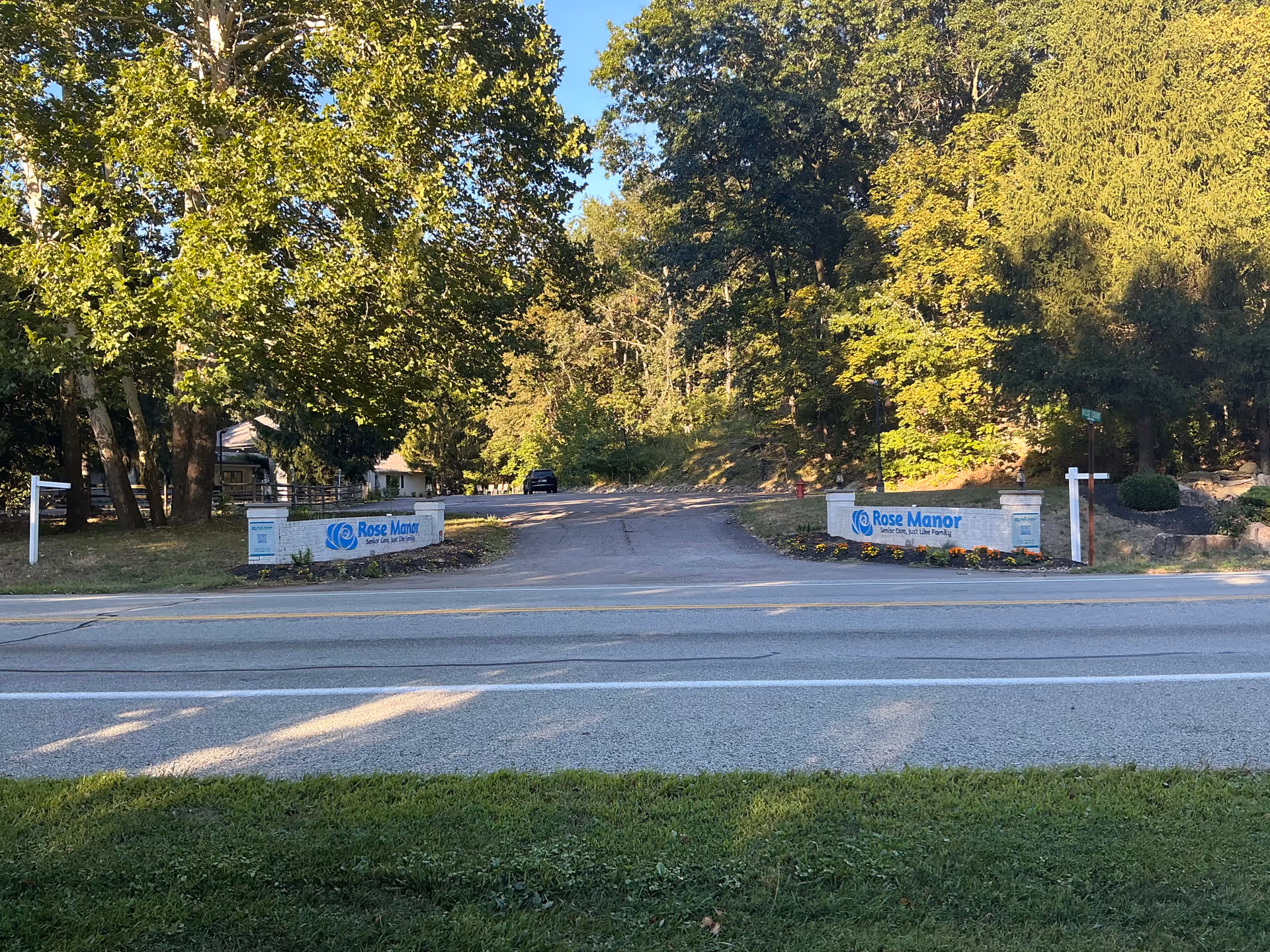 Entrance driveway to Rose Manor senior living facility with low brick signs and trees lining the road.