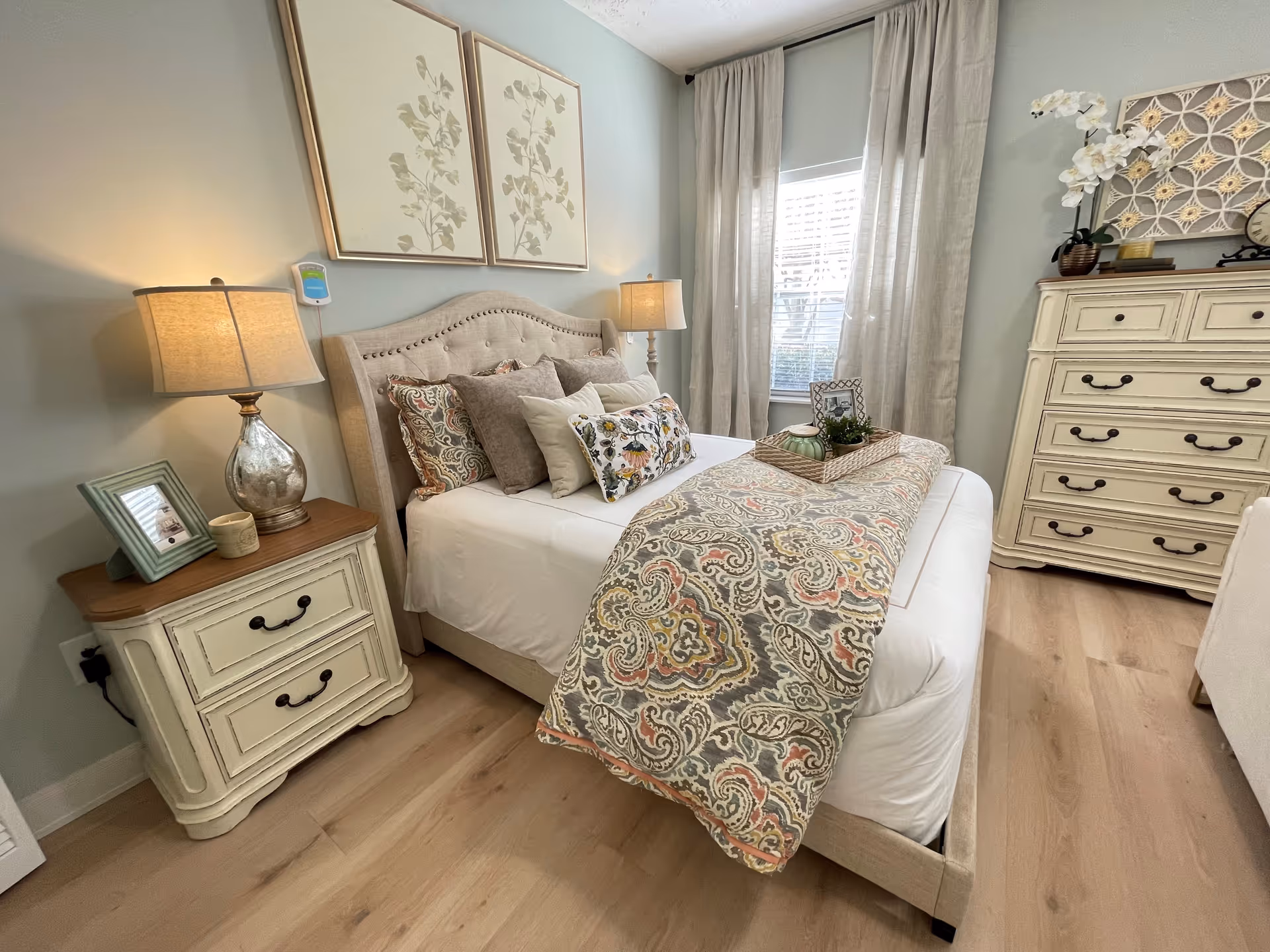 A cozy bedroom with a beige upholstered bed adorned with multiple decorative pillows and a patterned throw blanket. On the left side, there is a cream-colored nightstand with two drawers, a table lamp, a framed photo, and a small candle. On the right side, a matching cream-colored dresser with multiple drawers is decorated with a potted orchid, a clock, and a decorative wall piece above it. Light wood flooring and a window with beige curtains complete the room.