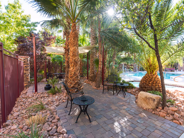 Outdoor patio area with stone pavers, metal chairs and tables, surrounded by palm trees and other greenery, next to a swimming pool with clear blue water under bright sunlight.