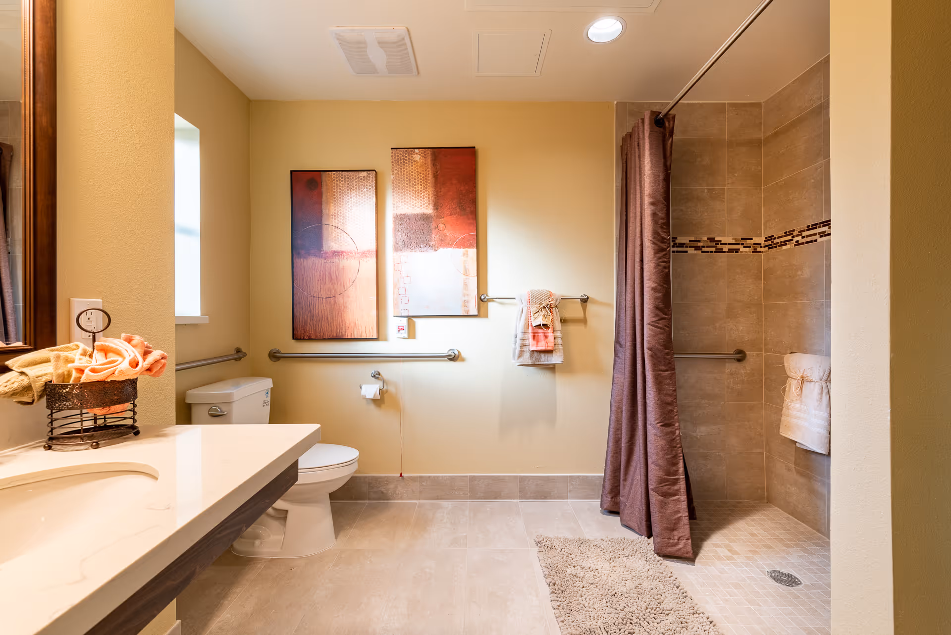 A spacious bathroom with beige tiled floor and walls. There is a white toilet next to a window, a countertop with a sink and towels on the left, and a walk-in shower with a brown curtain and tiled walls on the right. Two abstract paintings hang on the wall above the toilet, and there are grab bars installed near the toilet and inside the shower. A beige bath mat is placed in front of the shower.