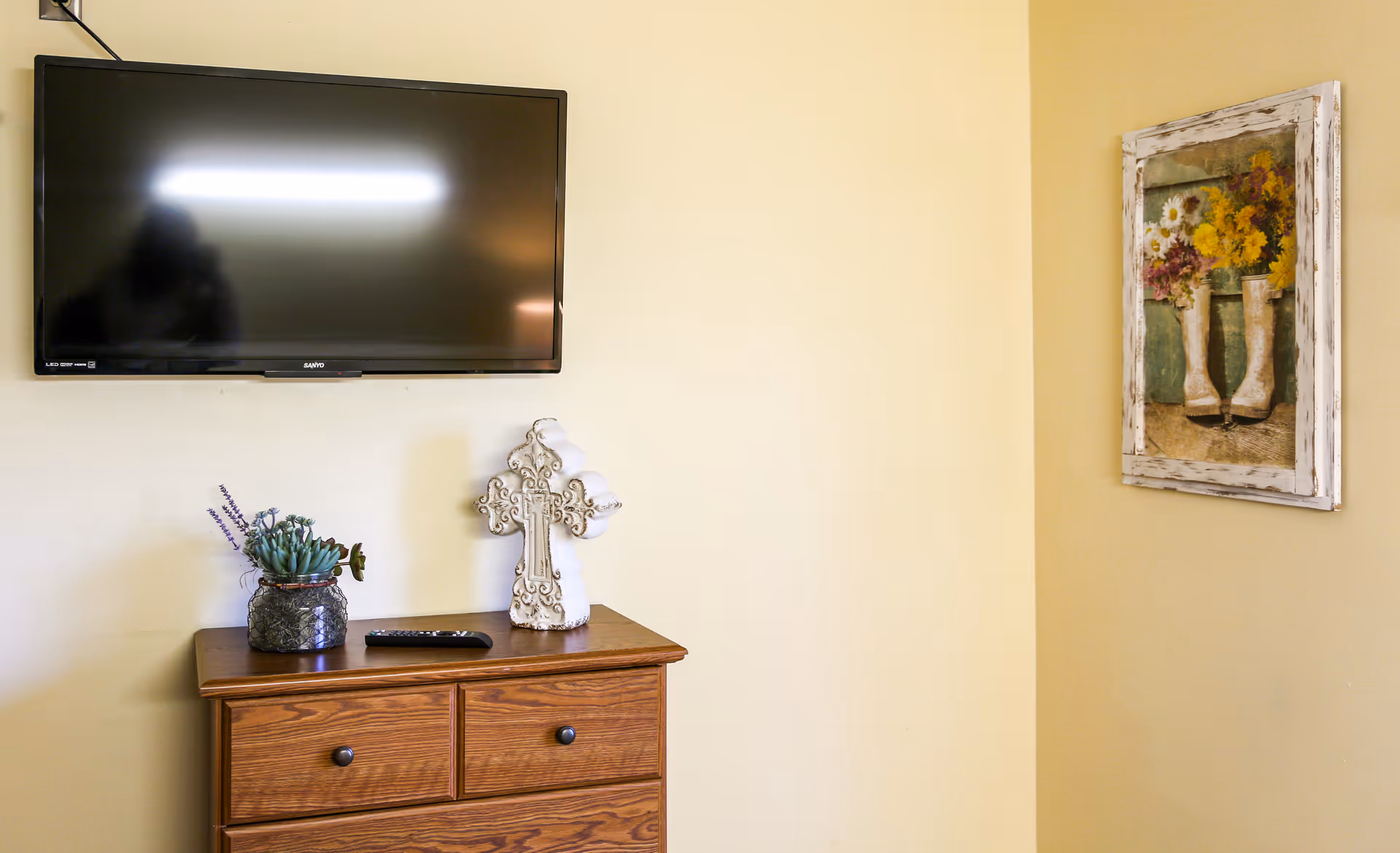 A wall-mounted flat screen TV above a wooden dresser with two drawers. On top of the dresser, there is a decorative cross, a small potted plant, and a TV remote. To the right on the wall, there is a framed painting of white boots with flowers inside them.
