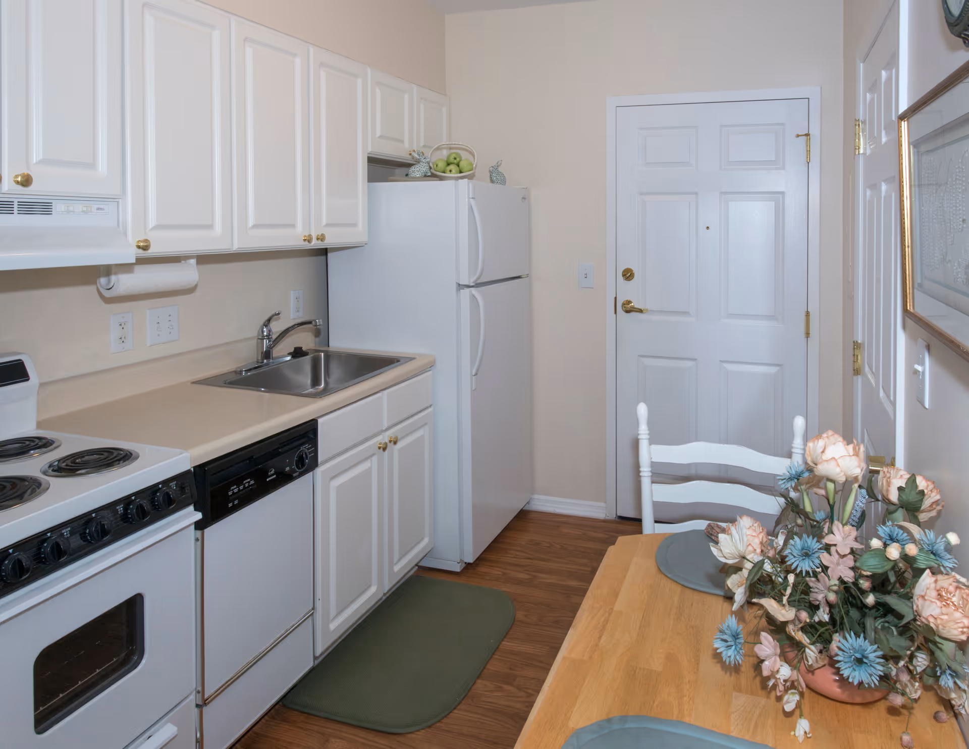 A compact kitchen area featuring white cabinets with gold knobs, a white refrigerator, a white stove with an oven, a dishwasher, and a stainless steel sink. There is a wooden dining table with two place settings and a floral centerpiece with pink and blue flowers. The floor is wood, and a green mat is placed in front of the sink. A white door is visible at the end of the kitchen.