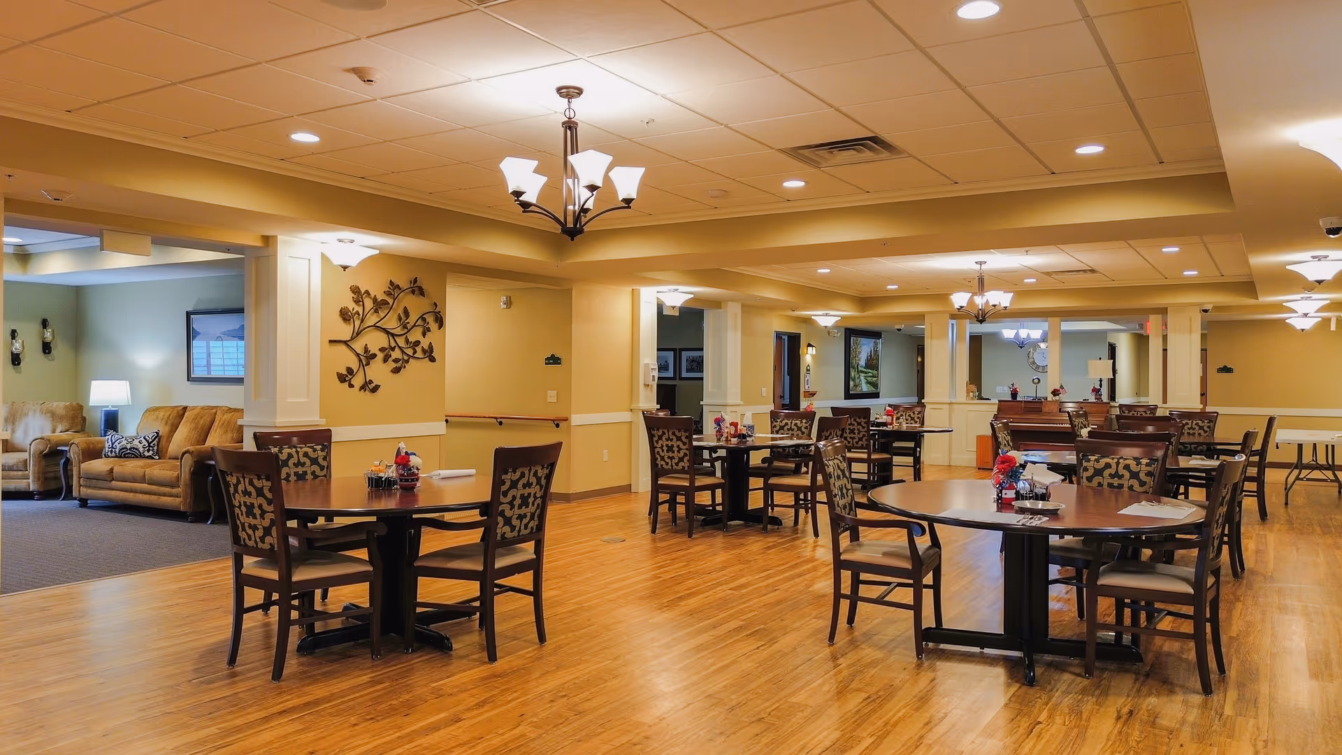 A spacious dining room in a senior living facility with multiple round wooden tables and chairs arranged neatly. The room has warm lighting with ceiling lights and chandeliers, wooden flooring, and beige walls. In the background, there is a seating area with sofas and a lamp, as well as decorative wall art and framed pictures.