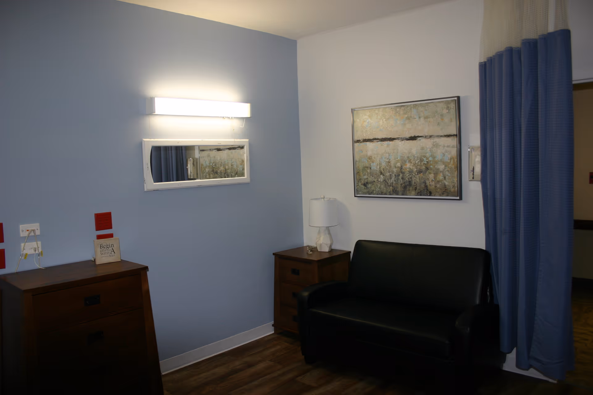A small room with light blue and white walls featuring a black leather loveseat, two wooden dressers, a table lamp, a rectangular mirror, and a framed abstract painting. A blue curtain is partially drawn to the right side of the room.