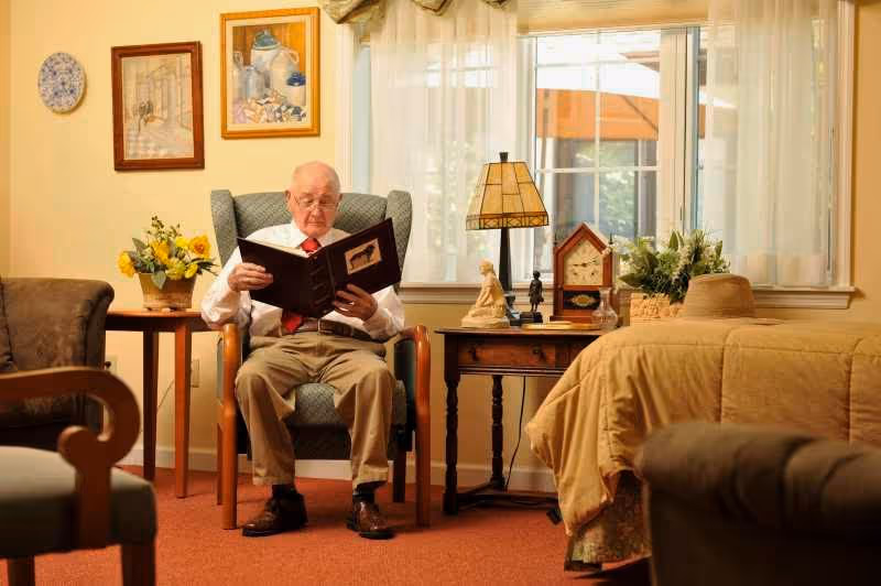 An elderly man sits in a chair reading a book in a warmly decorated assisted living bedroom with a bed, side tables, and a window.