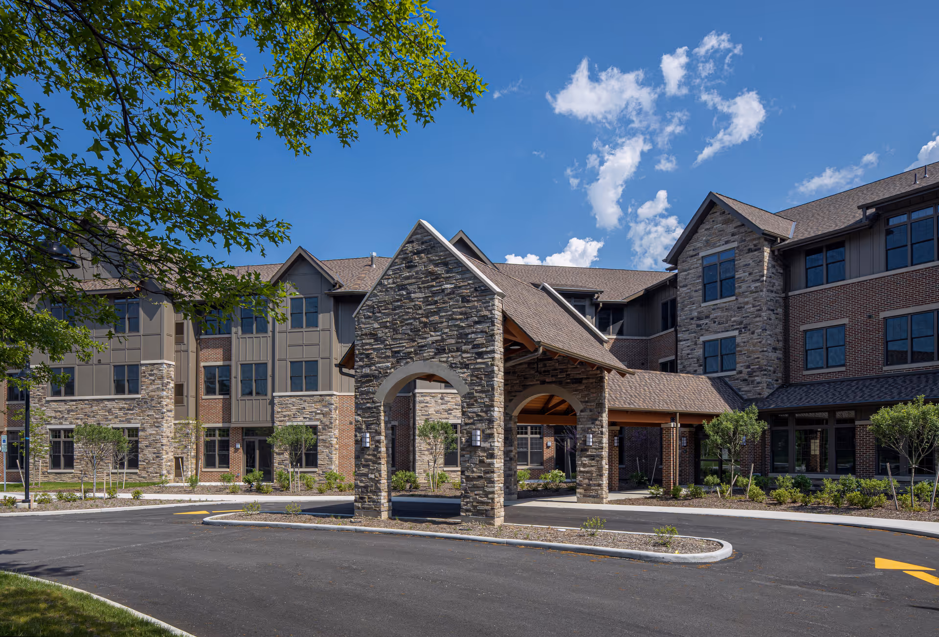 Exterior view of a senior living facility named Concord Reserve featuring a large stone and brick building with multiple windows, a covered entrance with stone pillars, landscaped greenery, and a clear blue sky with some clouds.