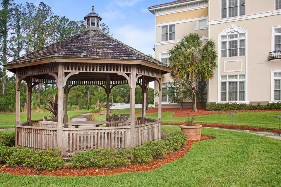 Wooden gazebo with benches on a landscaped lawn in front of a multi-story light-colored building.