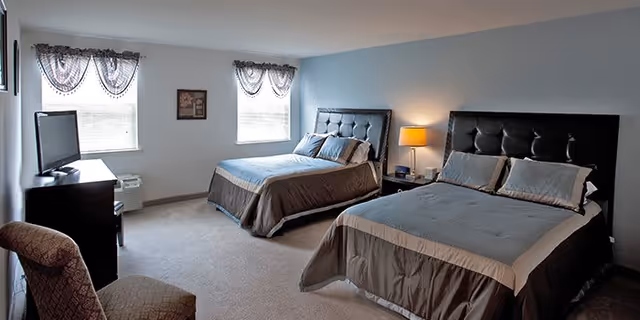 A bedroom with two large beds featuring dark tufted headboards and matching bedding. The room has light blue walls, two windows with sheer curtains, a small TV on a dark dresser, a patterned armchair, and a lamp on a nightstand between the beds.