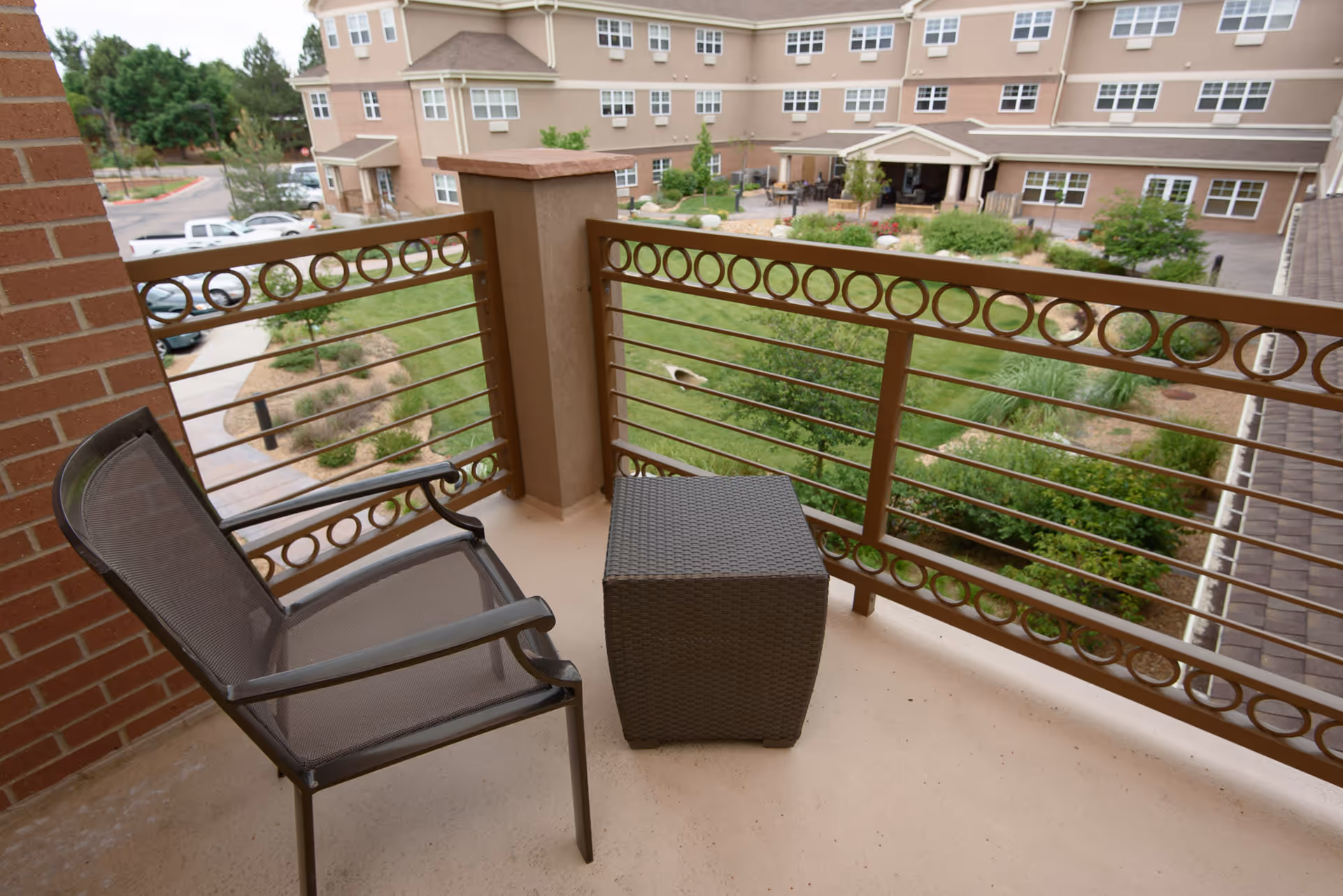 A small balcony with a metal chair and a woven square table overlooking a landscaped garden and a multi-story residential building in the background.