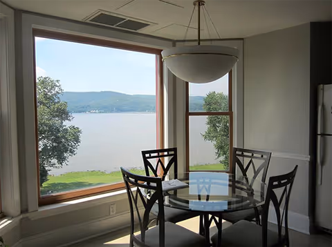 Dining area with a round glass table and four black chairs, large windows offering a scenic view of a lake, trees, and distant hills, and a hanging light fixture above the table.