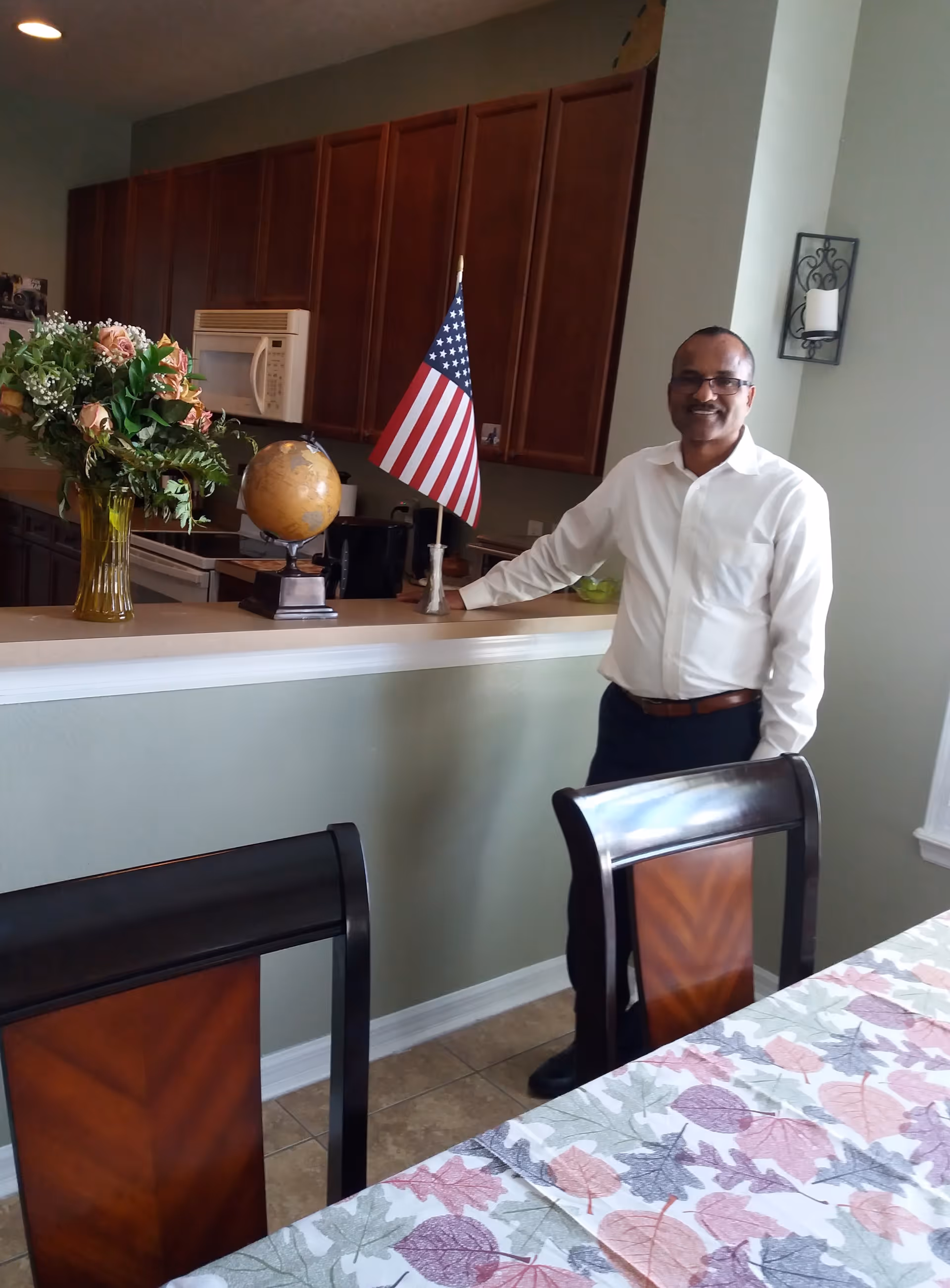 A man in a white shirt and dark pants stands smiling behind a kitchen counter. On the counter are a vase with flowers, a globe, and a small American flag. Behind him are wooden kitchen cabinets and a microwave. In the foreground, part of a dining table with a leaf-patterned tablecloth and two chairs is visible.