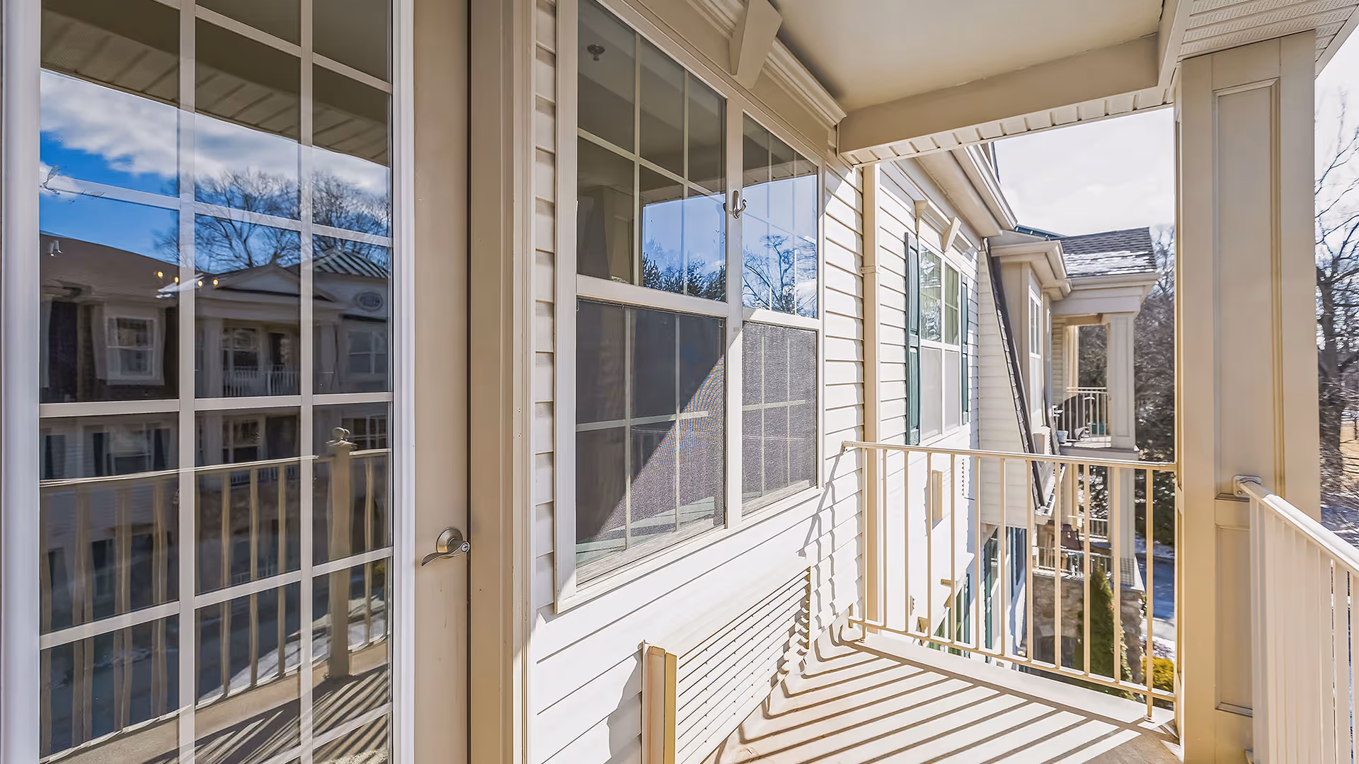 A bright outdoor balcony area with white railings and beige siding on the building. The balcony overlooks neighboring buildings and trees under a clear blue sky.