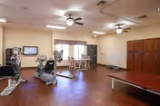 A spacious room with exercise equipment including stationary bikes and a treadmill, a table with chairs near a window, ceiling fans, and wooden cabinets along one wall. The room has a wooden floor and beige walls.