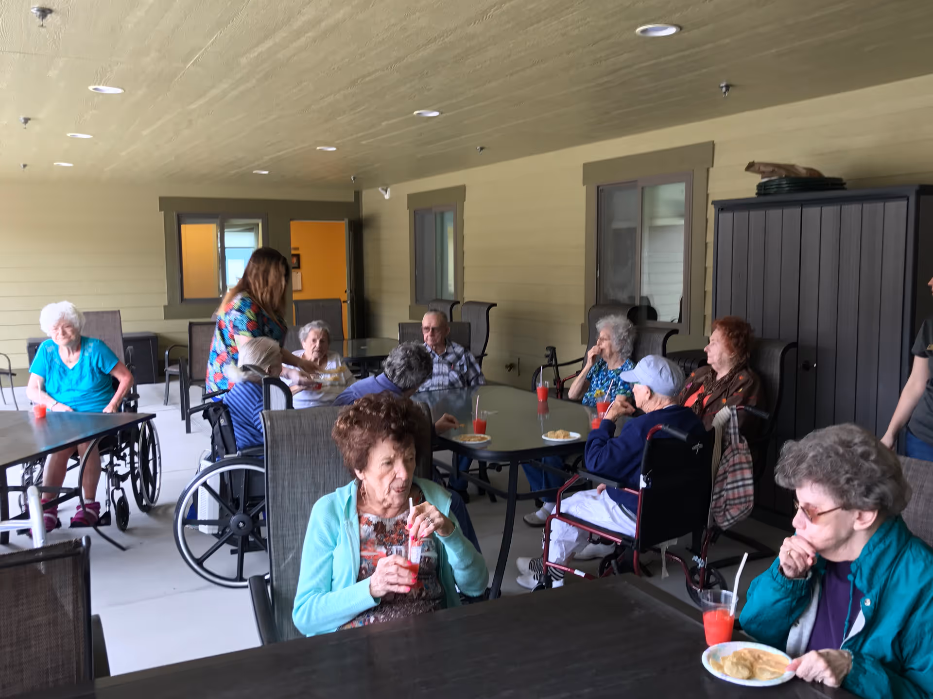 A group of elderly people sitting around tables on a covered outdoor patio area. Some are in wheelchairs, and they are drinking red beverages and eating snacks. A caregiver is assisting one of the residents. The setting appears to be a social or recreational gathering at a senior living facility.