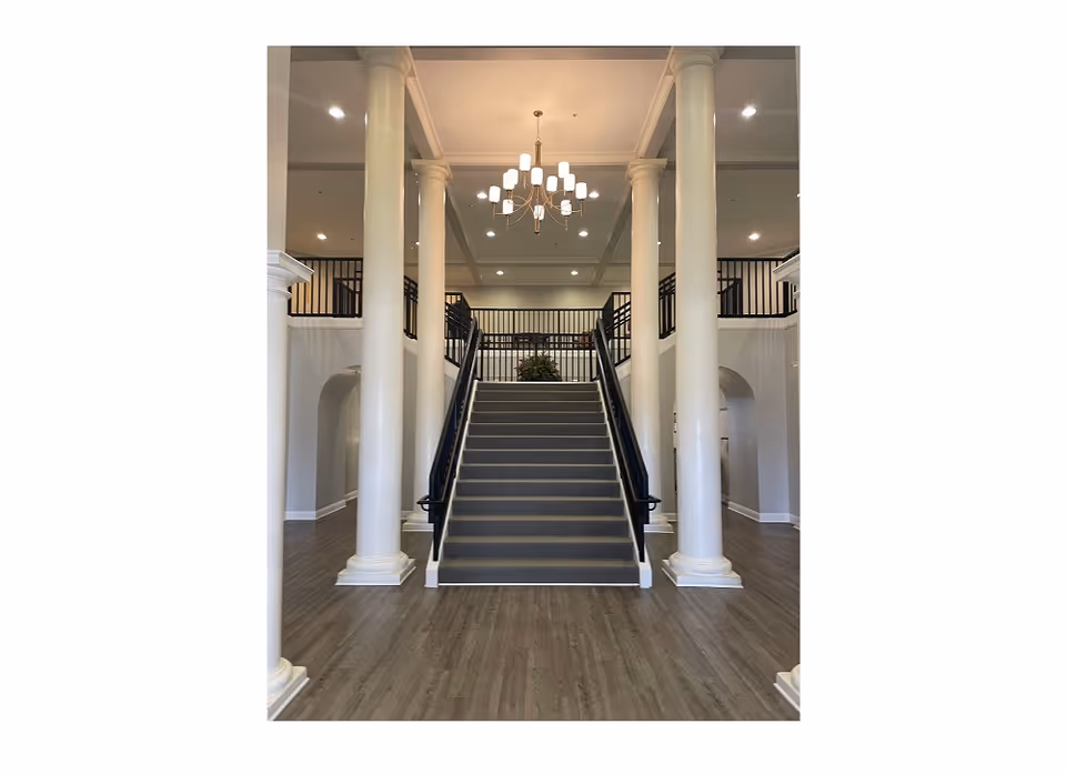Grand interior lobby with a central carpeted staircase flanked by tall white columns and a chandelier overhead.