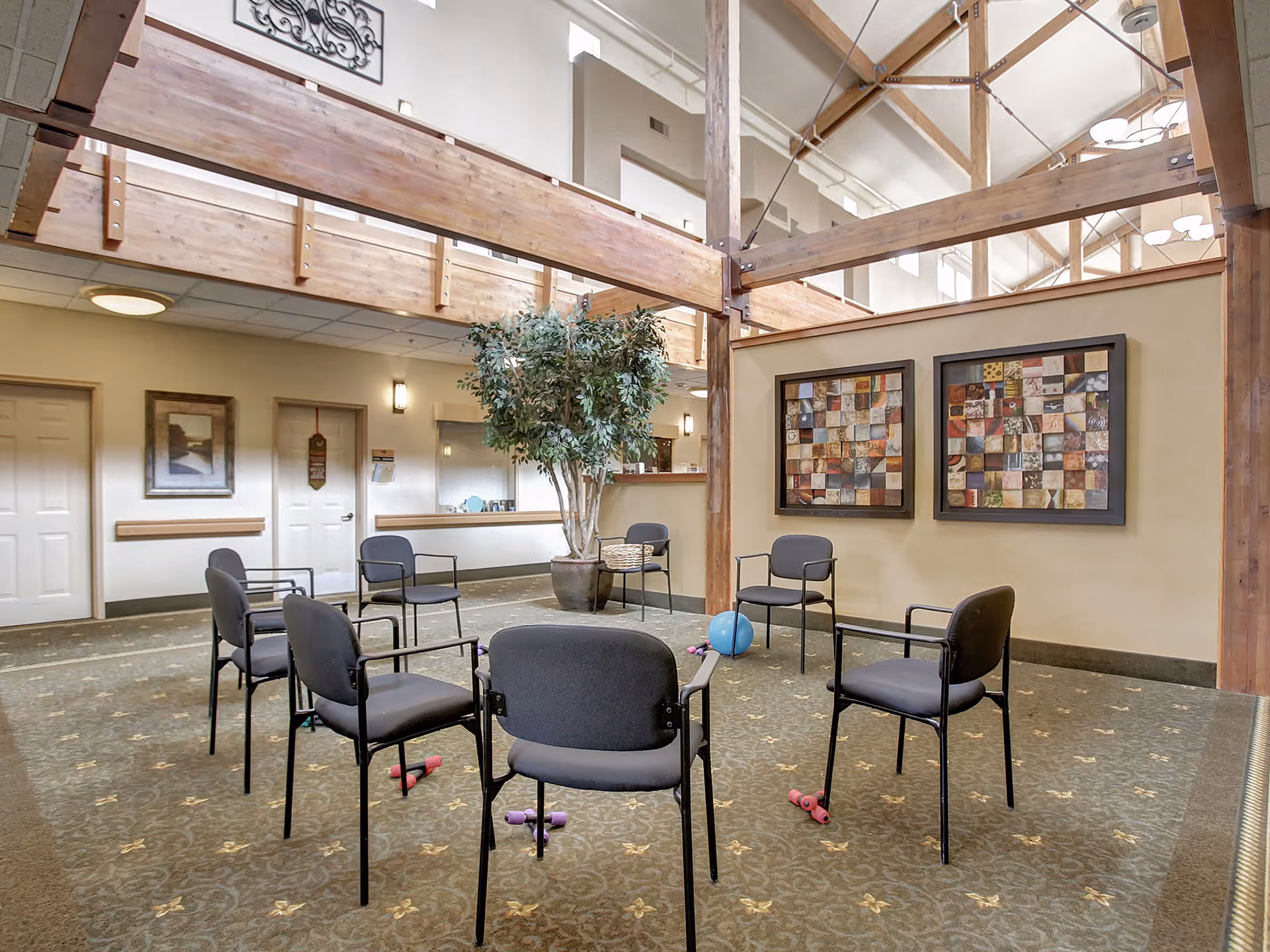 A spacious senior living common area with a circle of eight black chairs arranged on a patterned carpet. Small exercise weights and a blue exercise ball are placed on the floor inside the circle. The room features wooden beams, a potted tree, two framed abstract art pieces on the wall, and a reception window in the background.