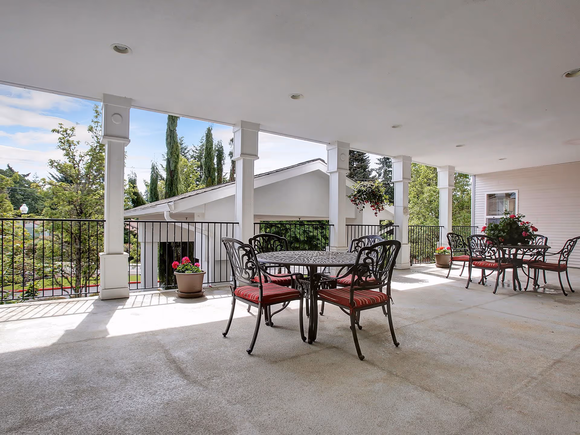 Covered outdoor patio area with two round metal tables and several chairs with red cushions. There are potted plants and hanging flower baskets around the patio, with a view of trees and a neighboring building in the background.