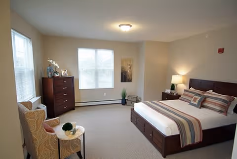 A spacious bedroom with a large bed featuring striped bedding and multiple pillows. There is a wooden dresser with decorative items and a framed photo on top, a comfortable armchair with a small round table holding a plant, and a lamp on a nightstand next to the bed. The room has beige walls, carpeted floor, and two windows letting in natural light.
