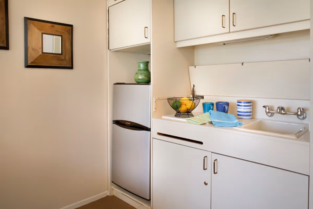 Small kitchenette area with white cabinets, a compact refrigerator, a sink with a wall-mounted faucet, a wire fruit basket containing lemons and limes, three colorful mugs, and folded dish towels. A small green vase is placed on top of the refrigerator. A wooden framed mirror hangs on the adjacent wall.