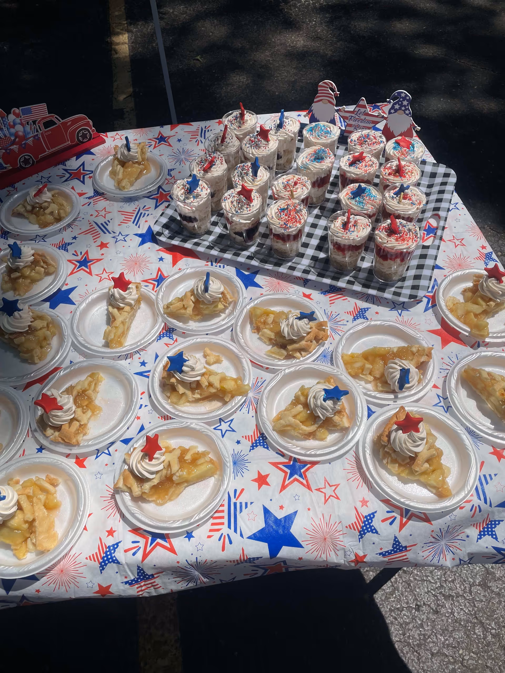 Outdoor table with plated slices of pie and cups of layered red-white-blue desserts on a patriotic star-patterned tablecloth.