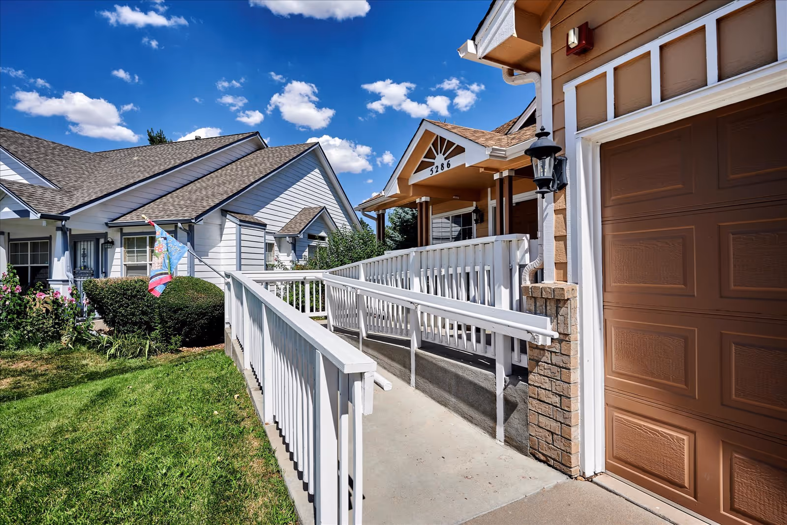 Exterior view of a senior living facility entrance with a white wheelchair ramp leading to a porch. The building has a brown garage door and beige siding with a house number 5286 above the porch. There is a green lawn and bushes on the left side, and a bright blue sky with scattered clouds overhead.