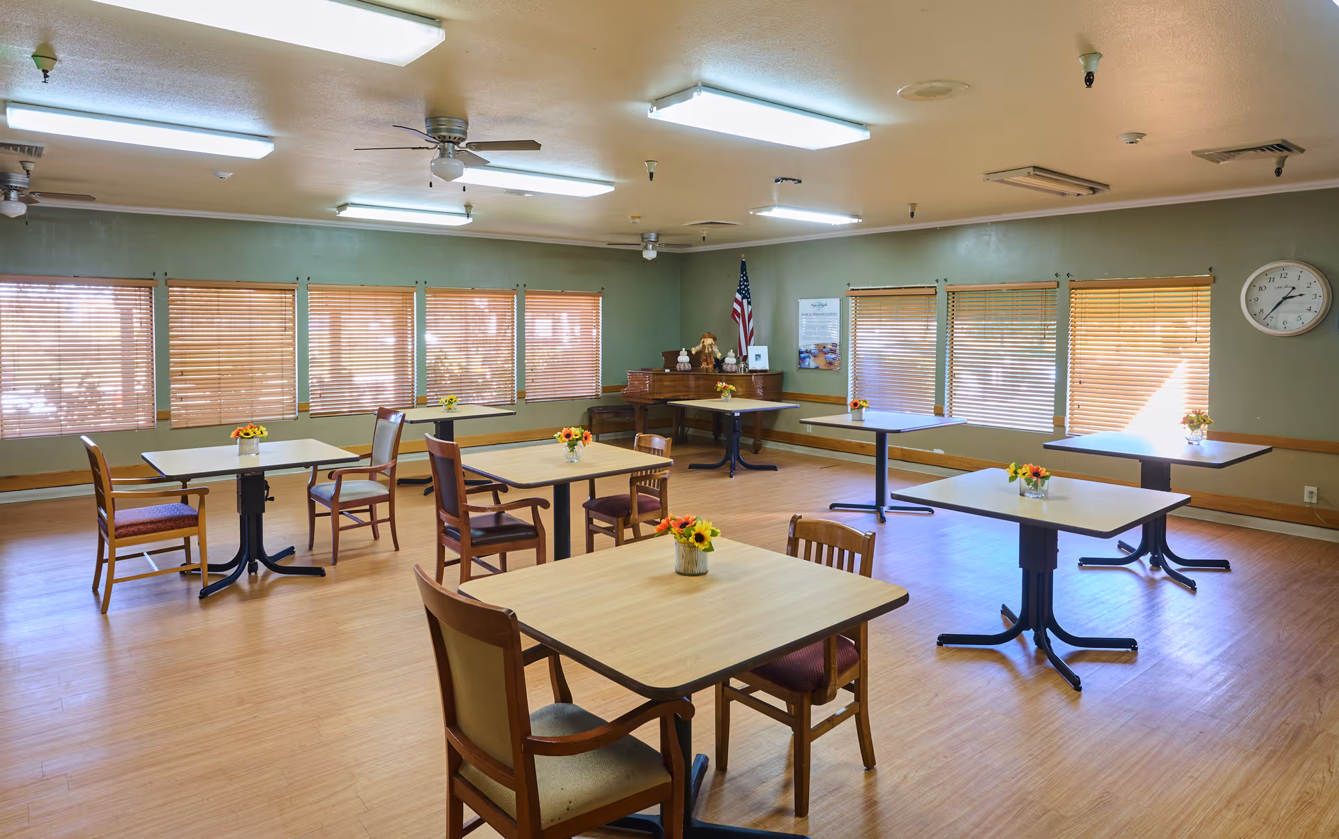 Communal dining room with several square tables and chairs, wooden blinds on the windows, and small floral centerpieces.