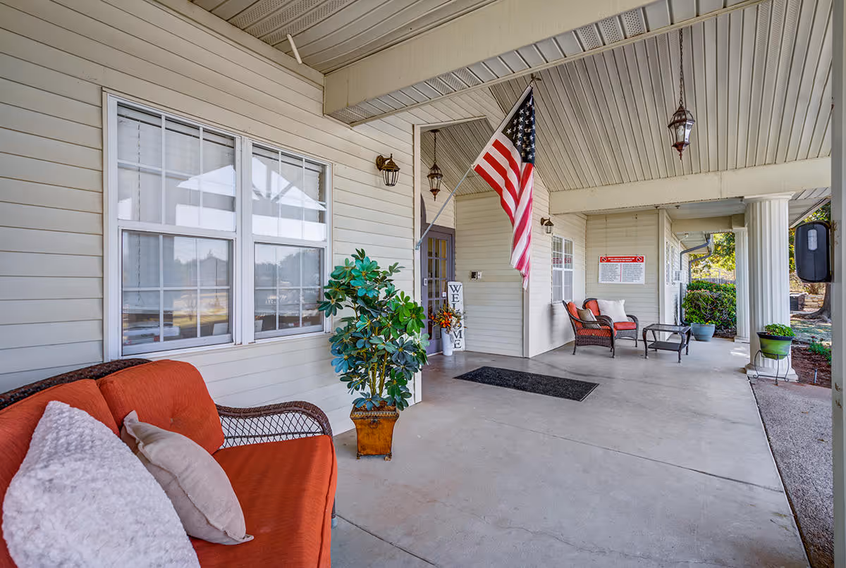 Covered outdoor patio area with beige siding walls, a ceiling with recessed lighting, and an American flag hanging from a pole. There are two seating areas with wicker furniture and red cushions, a potted plant, a welcome sign near the door, and a black doormat on the concrete floor.