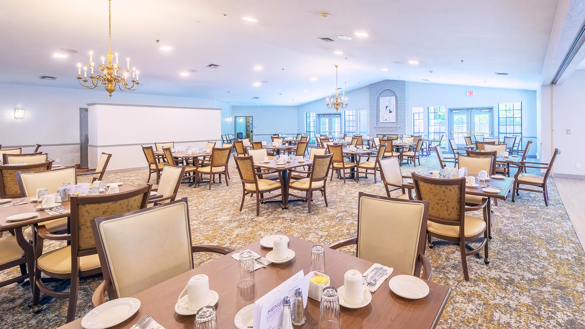 A spacious dining room with multiple wooden tables and cushioned chairs arranged neatly. Each table is set with white cups, plates, glasses, and silverware. The room features chandeliers hanging from the ceiling, large windows allowing natural light, and a carpeted floor with a patterned design. The walls are light-colored, and there is a fireplace with artwork above it in the background.